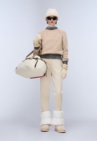 Knit sweater in beige with a textured finish, cream beige pants, white puffy boot cuffs, and a light-colored duffel bag with straps.
