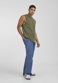 Top senza maniche verde oliva abbinato a jeans dritti azzurri. La modella è in piedi con una mano nella tasca, indossando sneakers grigie e bianche.