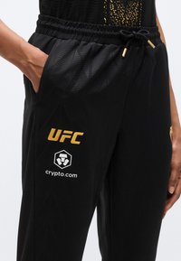 Pantalon de sport noir avec un motif texturé, une ceinture élastique et des poches avant. Orné de logos dorés "UFC" et "crypto.com".