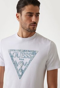 Balta medvilninė marškinėliai su mėlynu trikampio grafiniu dizainu, papuoštu nėriniuota tekstūra, ir centriniu "GUESS" logotipu. Apvalus kaklas.