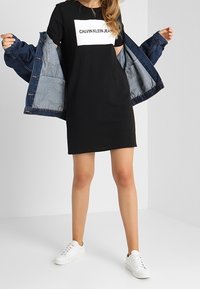 Černé tričkové šaty s bílým logem Calvin Klein. V kombinaci s modrou denimovou bundičkou a bílými teniskami, což podtrhuje ležérní styl.