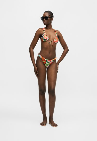 Γυναίκα με φλοράλ μπικίνι με βολάν και γυαλιά ηλίου, όρθια ξυπόλυτη σε απλό λευκό φόντο.