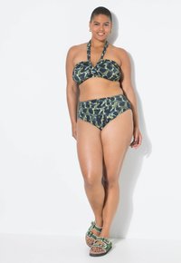 Zelené a černé vzorované bikiny s halter topem a vysokým pasem. Modelka má na sobě zelené sandály s bílými akcenty. Bílý pozadí.