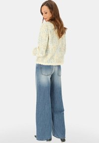 Kobieta w kremowej, fakturowanej kurtce i szerokich, wyblakłych niebieskich dżinsach, stojąca tyłem.