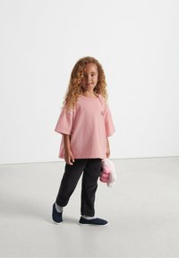 Lyserød oversized t-shirt, ribbede sorte bukser og blå slip-on sko. Barn holder et blødt lyserødt stuffed dyr. Krøllet hår.