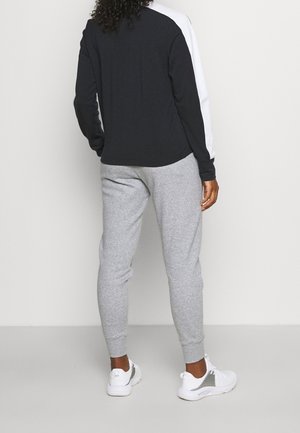 Person iført en sort og hvid langærmet top, lysegrå joggingbukser og hvide sneakers, vendt væk fra kameraet mod en ensfarvet baggrund.