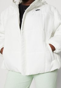 White puffer jacket with a zip front, elastic cuffs, and pockets. Features a subtle black Nike logo on the chest. Soft, padded texture.