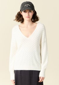 Maglione bianco lavorato a maglia con un motivo a diamante, scollatura a V profonda e maniche lunghe. Indossato con un berretto nero con ricami bianchi.