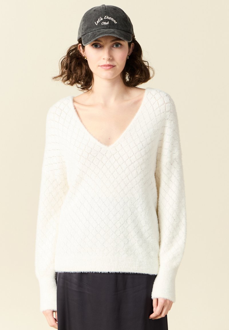 Maglione bianco lavorato a maglia con un motivo a diamante, scollatura a V profonda e maniche lunghe. Indossato con un berretto nero con ricami bianchi.
