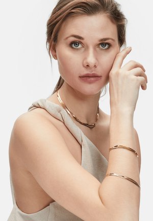Rozen goud sieraden omvatten een gebogen choker en bijpassende armbanden, waarbij een gladde, reflecterende oppervlakte en een minimalistisch ontwerp te zien zijn tegen neutrale stoffen.