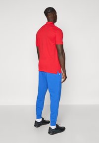 Red short-sleeve polo shirt with side slits, paired with blue jogger pants featuring elastic cuffs, worn with black athletic shoes.