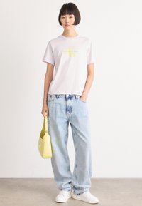 Lys rosa bomull t-skjorte med grønn og gul Calvin Klein-logo, kombinert med løse, lyseblå denimjeans og hvite joggesko. Holder en gul veske.