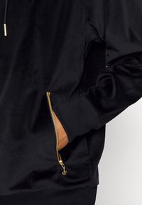 Svart velourhoodie med en guldlåsficka och ribbade muddar. Tyget har en slät textur som framhäver den eleganta designen.