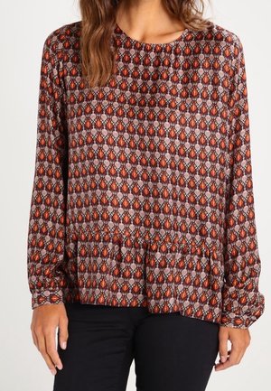 Kobieta ubrana w bluzkę z długim rękawem o geometrycznym wzorze w kolorach brązu, pomarańczy i czerni, w połączeniu z czarnymi spodniami.