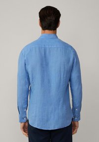 Camicia a maniche lunghe di lino di colore blu chiaro, con colletto e polsini a bottoni, orlo leggermente curvo e texture liscia.