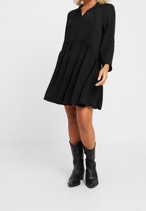 Vrouw draagt een zwarte, lange mouw, los vallende jurk tot op de knie, gecombineerd met zwarte leren laarzen tot halverwege de kuit, voor een effen achtergrond.