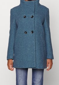 Zeleno-modrý vlna smíšený kabát s vysokým límcem, dvouřadým zapínáním a černými knoflíky. Má hladkou texturu a boční kapsy.
