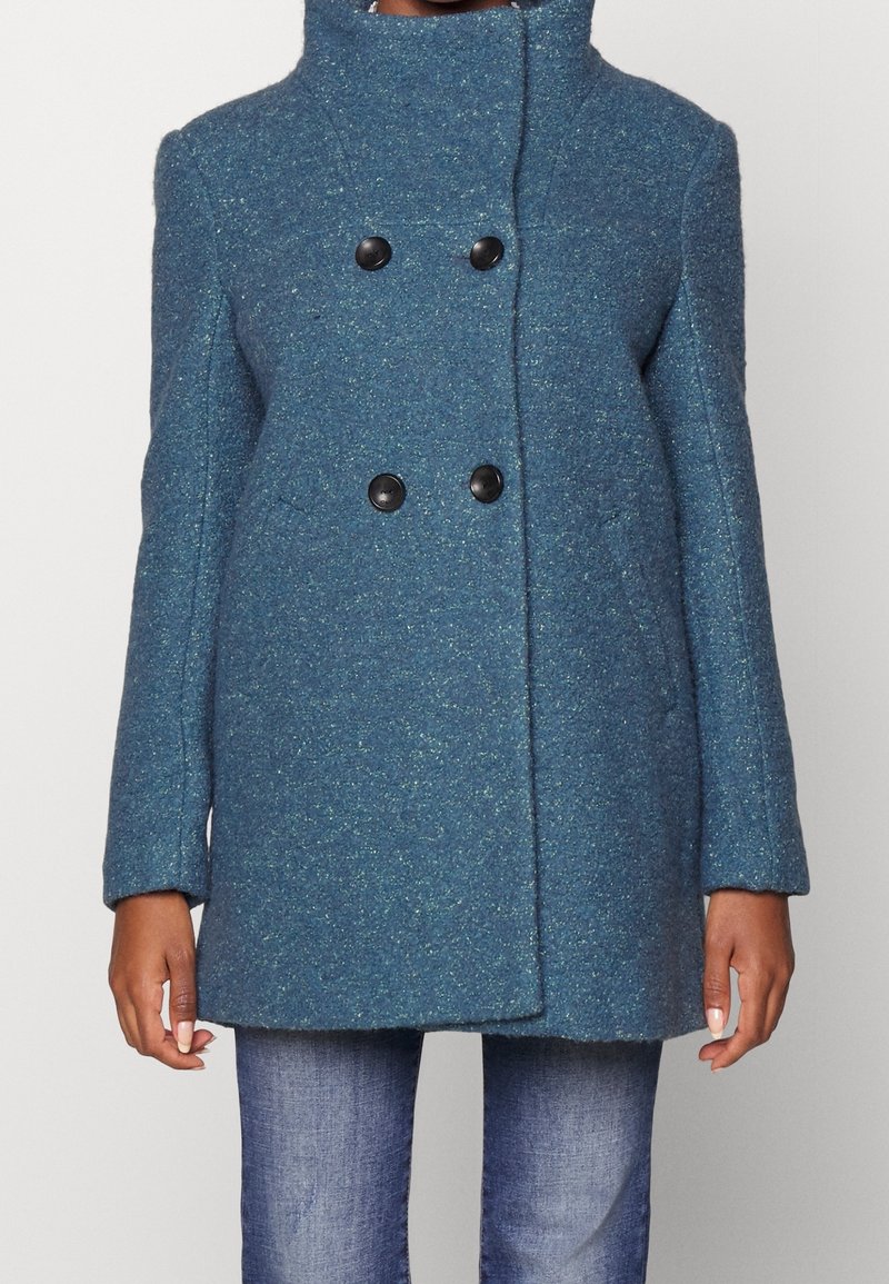 Zeleno-modrý vlna smíšený kabát s vysokým límcem, dvouřadým zapínáním a černými knoflíky. Má hladkou texturu a boční kapsy.
