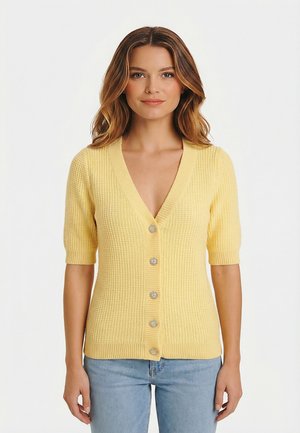 Donna che indossa un cardigan lavorato a maglia a maniche corte di colore giallo pallido con cinque bottoni decorativi, abbinato a jeans azzurri, in piedi davanti a uno sfondo bianco semplice.
