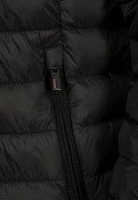 Czarna kurtka puchowa o teksturowanym, pikowanym wzorze z wyraźnym zamkiem. Metka z logo widoczna w pobliżu zamka w celu identyfikacji.