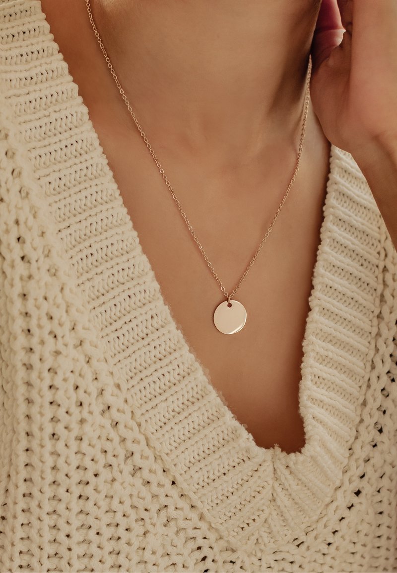 Rosenvergoldete, runde Anhänger-Halskette an feiner Kette, präsentiert vor einem strukturierten cremfarbenen Strickpullover mit weitem Ausschnitt.