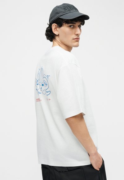 Νεαρός άνδρας που φοράει λευκό T-shirt με μπλε γραφικό στην πλάτη, μαύρο καπέλο, σκουλαρίκια και μαύρο παντελόνι, κοιτάζει πάνω από τον ώμο του με φόντο λευκό.