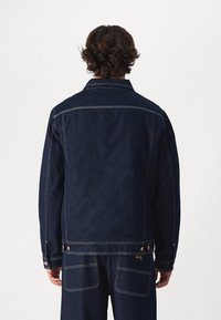 Tmavě modrá džínová bunda s klasickým límečkem, viditelným prošitím a kovovými doplňky v barvě mědi. Má rovný střih a kapsy na zádech.