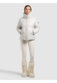 Donna che indossa una giacca imbottita bianca, leggings color crema e stivali invernali beige, in piedi davanti a uno sfondo grigio chiaro.