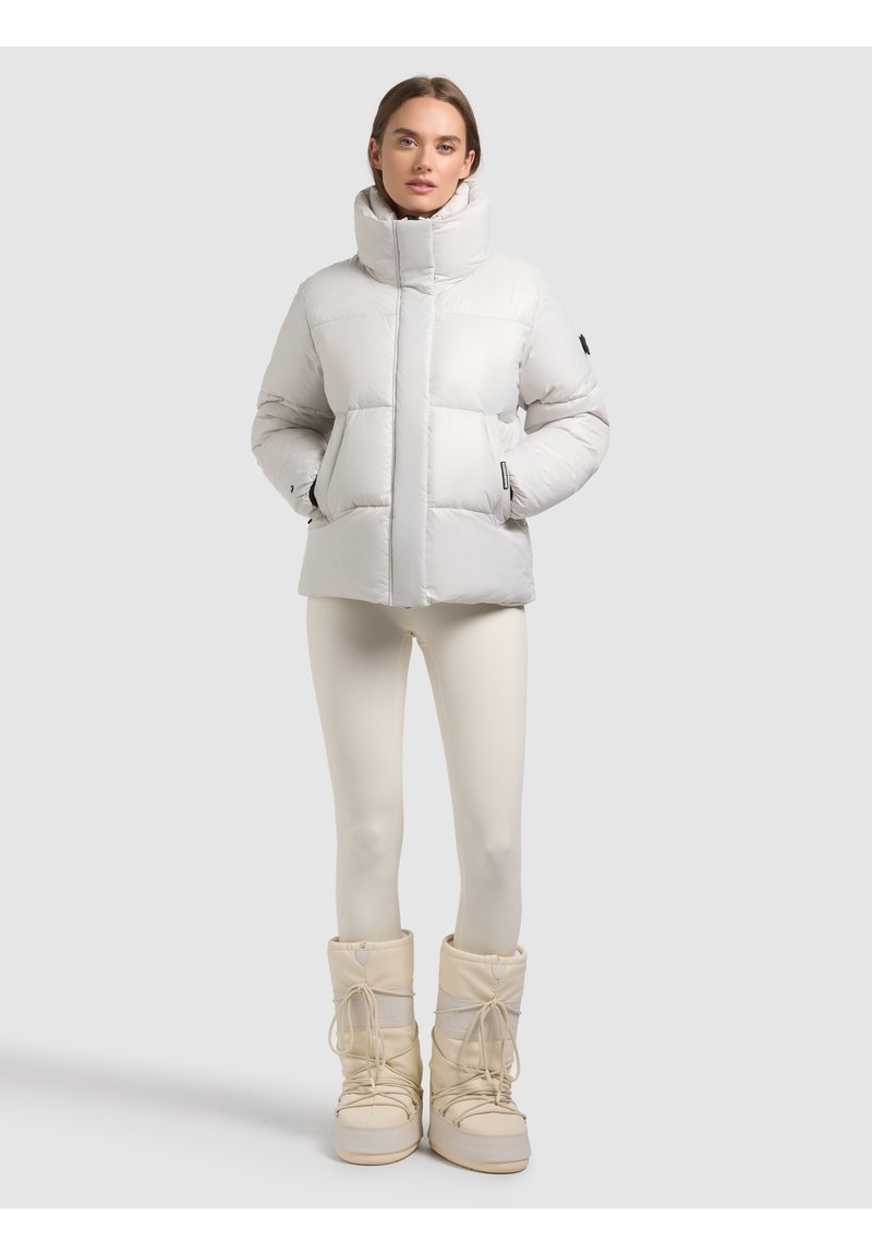 Donna che indossa una giacca imbottita bianca, leggings color crema e stivali invernali beige, in piedi davanti a uno sfondo grigio chiaro.