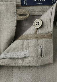 Gros plan sur un pantalon en lin beige montrant le bouton, la boutonnière, la boucle de la ceinture et la braguette partiellement ouverte avec des coutures visibles.