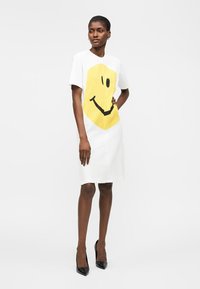 Nő állva, fehér térdig érő ruhát visel rövid ujjakkal és nagy sárga mosolygós arc grafikával, fekete hegyes sarkú cipővel párosítva.