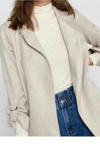 Cappotto in suede beige con bavero e maniche arrotolate, abbinato a un dolcevita crema e jeans in denim blu a vita alta. Leggera texture e vestibilità rilassata.