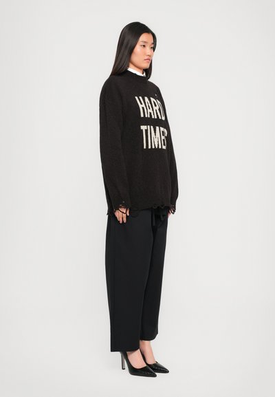 Black oversized sweater with "HARD TIMES" in white text, paired with black tailored trousers and black patent heels.