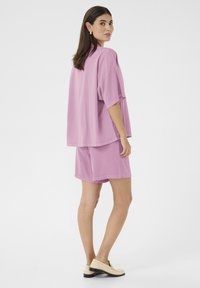 Rozā pārsteidzīgi t-krekls un atbilstošu šortu komplekts. Mīksta, gluda auduma ar smalku tekstūru. Modelis stāv ar muguru griezusies, uzvilkusi gaišas kurpes.