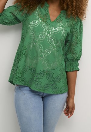 Frau trägt grüne Spitzenbluse mit floralem Muster und dreiviertel Puffärmeln, kombiniert mit hellblauen Jeans.