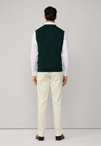 Granatowa zielona kamizelka z dzianiny na białej koszuli z długim rękawem, zestawiona z kremowymi spodniami i czarnymi butami, widok z tyłu.