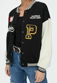 Czarna i biała kurtka varsity z haftowanymi naszywkami, prążkowanymi mankietami i kołnierzem, z zapięciem na guziki oraz kieszeniami bocznymi. Nosi się ją na czarnym topie.