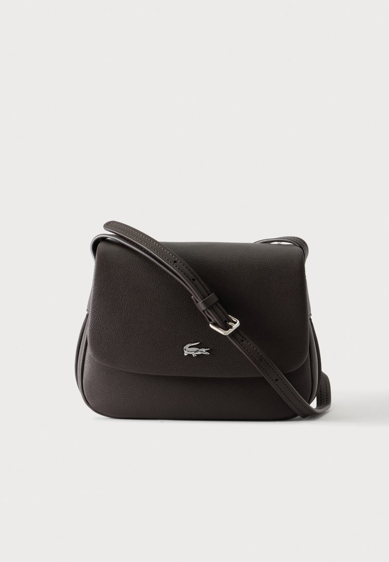 Borsa a tracolla in pelle nera con tracolla regolabile e piccolo logo di coccodrillo argentato sulla patta anteriore su uno sfondo chiaro uniforme.
