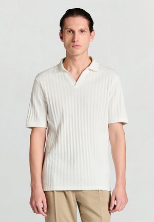 Polo blanc côtelé avec col en V, manches courtes et col doux. Le tissu présente une texture apparente.