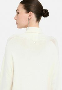 Maglione a maglia crema con colletto alto, finitura strutturata e un bottone decorativo sul retro. Maniche lunghe e vestibilità ampia.