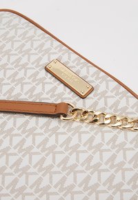 Biała i beżowa torba Michael Kors z powtarzającym się wzorem "MK", z brązowym skórzanym wykończeniem, złotym łańcuchem i prostokątną złotą plakietką z logo.
