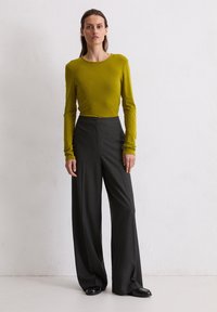 Langärmliges, olivgrünes Oberteil kombiniert mit hochtaillierten, grauen Nadelstreifenhosen. Schwarze flache Schuhe vervollständigen das Outfit.