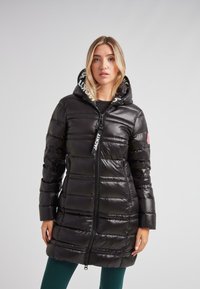 Mujer vistiendo un abrigo acolchado negro largo con capucha, bolsillos con cremallera y tirador de cremallera de marca, de pie contra un fondo claro y liso.