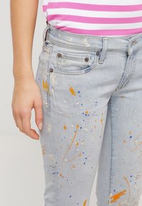 Mano izquierda junto a unos jeans azul claro con manchas de pintura azules, naranjas y blancas, combinados con una camiseta a rayas rosa y blanca.