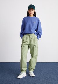 Ljusblå sweatshirt med en liten logotyp, gröna cargobyxor med sidofickor, vita och mintgröna sneakers samt en mörkgrön rutmönstrad keps.