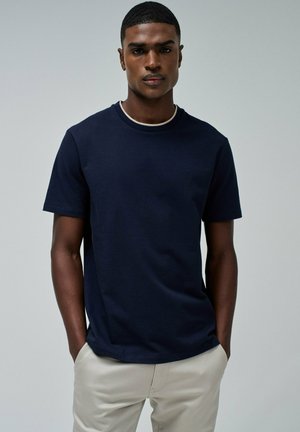 Camiseta de manga corta azul marino con cuello redondo y una textura sutil. El modelo lleva pantalones beige y tiene las manos en los bolsillos.