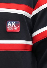 Camisa de rayas horizontales en azul marino y blanco, que presenta un parche bordado rectangular que dice "AX 1991" en rojo y blanco.