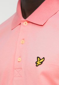 Ροζ πόλο μπλουζάκι με υφή στο ύφασμα, που διαθέτει κίτρινο κεντημένο λογότυπο ενός πουλιού στην αριστερή πλευρά και πορτοκαλί κουμπιά.