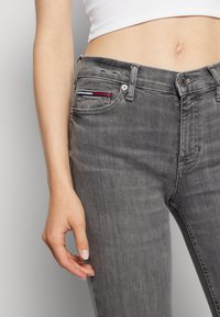 Jeans de mezclilla gris con un diseño ajustado, que cuentan con un pequeño bolsillo con rayas rojas, blancas y azules, y un cierre de botón. Textura suave.
