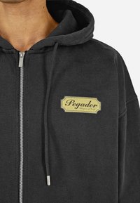 Juoda zip-up sudedama striukė, pagaminta iš minkštos medžiagos, su geltonu logo ženklu priekyje. Yra su raištelių gobtuvu ir sidabriniu užtrauktuku.